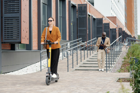 Young active businesswoman in eyeglasses and quiet luxury apparel riding electric scooter and moving along modern building of office centerの写真素材