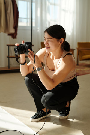 Young smiling female photographer with photocamera sitting on squats in front of camera while taking pictures of model during photo sessionの写真素材