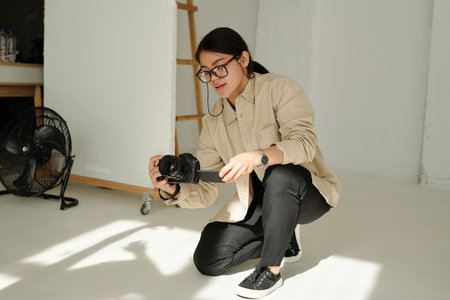 Brunette girl looking at screen of photocamera while standing on knee during photo session and taking new shot of model in studioの写真素材