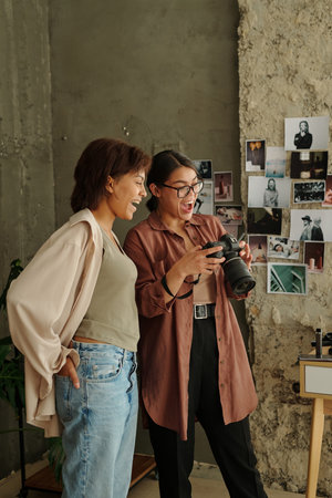 Cheerful young fashion model and photographer looking through new pictures on screen of photocamera taken during photo sessionの写真素材