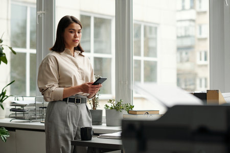 Young businesswoman in quiet luxury attire standing by office window and looking at printer with...