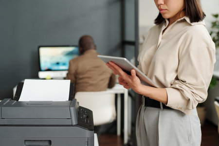 Focus on tablet held by young elegant businesswoman computing in office while standing by xerox...