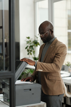 Serious African American male manager standing in front of xerox machine and putting paper on copier...