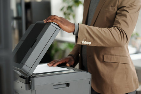 Cropped shot of young African American man putting paper on copier screen of xerox machine to make...
