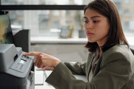 Young brunette businesswoman in grey blazer pressing button on panel of copier machine while...