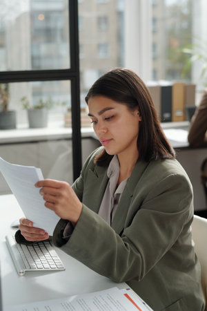 Young female secretary in formalwear looking through financial papers while preparing documents for...