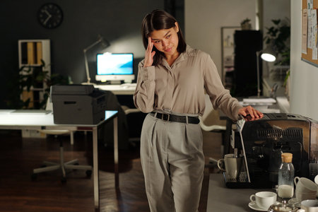 Young tired businesswoman in grey attire making cup of coffee in machine while standing against her...
