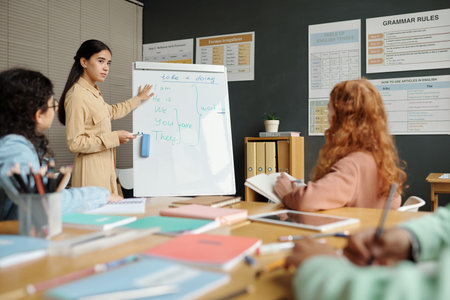 Young confident teacher standing by whiteboard with English grammar rules and pointing at one of...