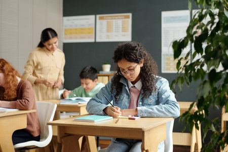 Diligent multiethnic schoolgirl making notes in copybook during individual work at lesson while...