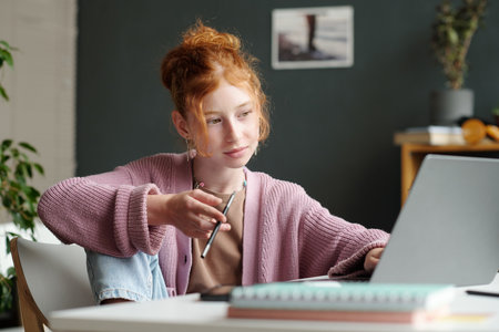 Serious schoolgirl in pink pullover listening to teacher during online lesson while sitting by desk...