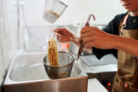 Young chef in apron putting hot boiled ramen into colander over metallic sink while standing in...