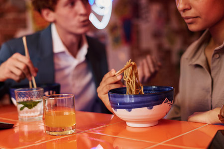 Focus on appetizing Japanese ramen on wooden chopsticks held by girl over bowl during lunch break with her colleague or boyfriend in cafeの写真素材