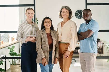 Group of successful intercultural white collar workers in smart casualwear looking at camera with smiles while standing in spacious officeの写真素材