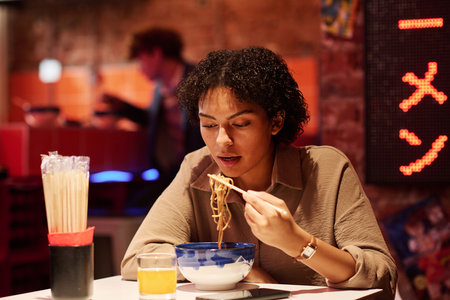 Young woman eating by table against hieroglyph meaning best ramen while sitting in cafe or restaurant selling traditional Japanese foodの写真素材