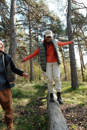 Happy young woman with her arms outstretched walking down dry log while enjoying autumn weekend with her husband or boyfriendの写真素材