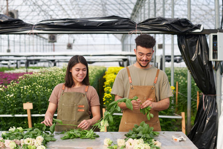 Two happy young multicultural florists in aprons sorting heap of fresh eustomas while standing by table with many flowers in greenhouseの写真素材