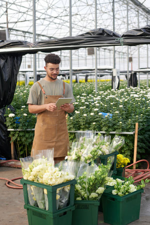 Young multiethnic male clerk of garden center or florist market looking at tablet screen while...