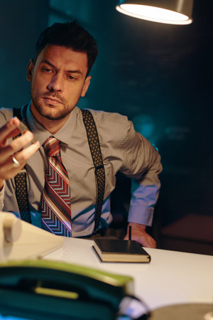 Young serious businessman in retro formalwear looking at mobile phone in his hand while sitting by workplace in front of camera in dark officeの写真素材