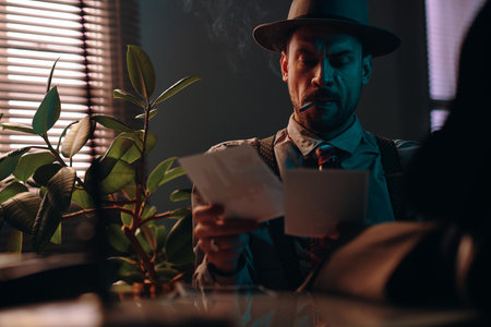 Young man with cigarette in mouth looking through photos and smoking while sitting in dark office or room with venetian blinds on windowの写真素材