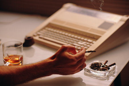 Hand of young male clerk shaking off ashes in ashtray and having glass of whiskey while sitting by workplace with old typewriterの写真素材