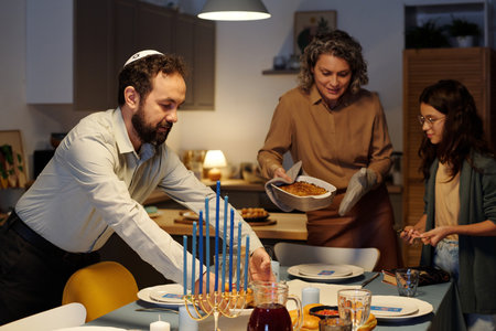 Bearded man with kippah on head putting plate with homemade snacks on table while serving it for guests with his wife and daughterの写真素材