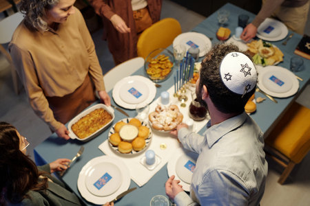 High angle of Jewish man wearing white kippah with embroidered star of David putting plate with homemade snack on served tableの写真素材
