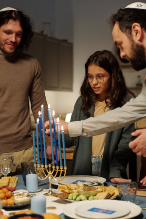 Mature man helping his daughter light candles of menorah while preparing for Hanukkah celebration by served tableの写真素材