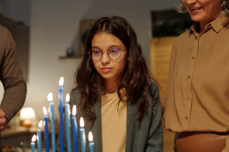 Adorable pre-teen girl in eyeglasses looking at burning blue candles on menorah candlestick while standing by her smiling motherの写真素材