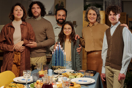 Family of six members of various generations standing by served table with burning menorah candles and homemade food and looking at youの写真素材