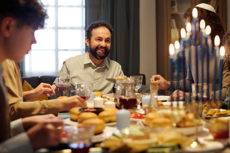 Cheerful mature man looking at one of guests during Hanukkah dinner with his family while enjoying homemade food and drinksの写真素材