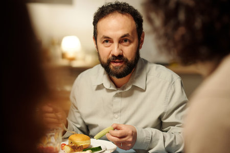 Mature Jewish man with piece of fresh cucumber in hand looking at guest or family member during chat by Hanukkah dinnerの写真素材