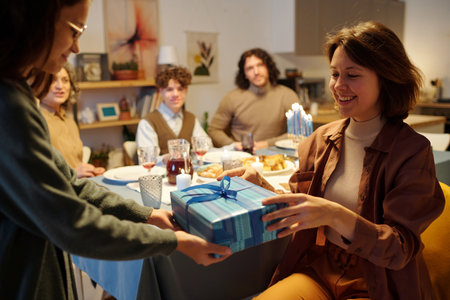 Young smiling woman taking blue giftbox with Hanukkah present from hands of her daughter against other members of family having dinnerの写真素材
