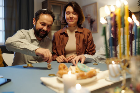 Smiling bearded man spinning small wooden dreidel on grey table with coins while sitting next to his daughter or wife during leisure gameの写真素材