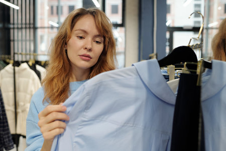 Young blond female consumer looking at blue cotton shirt from new seasonal fashion collection while choosing clothes in boutiqueの写真素材