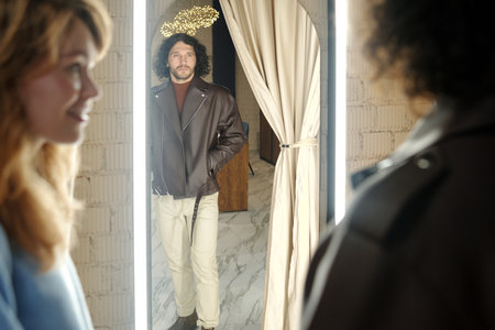 Reflection in mirror of young man trying on stylish leather jacket in fitting room in boutique and consulting with his wife or girlfriendの写真素材