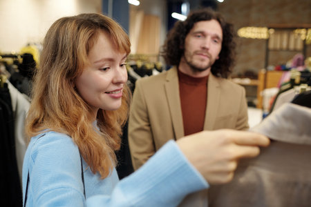 Young blond woman in blue pullover looking at coat on hanger while choosing new attire from seasonal collection with her husbandの写真素材