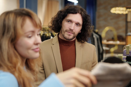 Young serious man in elegant quiet luxury attire looking at his wife or girlfriend choosing new jacket or coat from seasonal collectionの写真素材