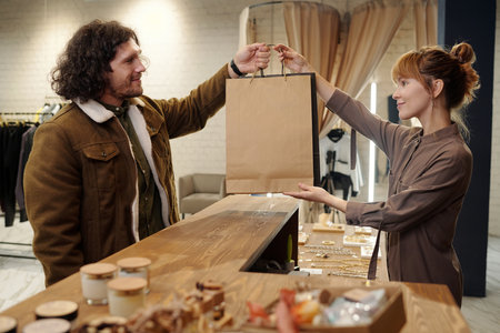 Young female shop assistant passing paperbags with purchases to male client of boutique over wooden counter after paying for stuffの写真素材