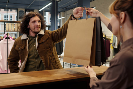 Young man in warm brown jacket taking paperbags from hands of female shop assistant while standing by counter in boutique showroomの写真素材