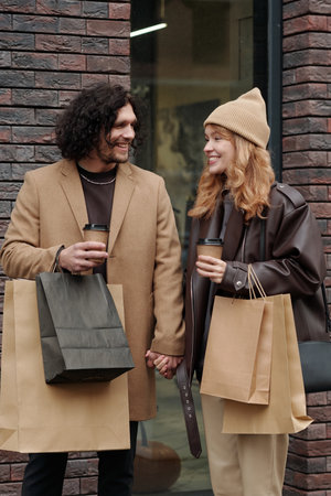 Young smiling man and woman with takeaway coffee and paperbags looking at one another while standing by brick buildingの写真素材