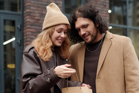 Pretty woman in stylish casualwear opening paperbag with purchase and boasting to her boyfriend in urban environment after shoppingの写真素材