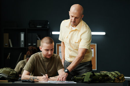 Confident mature coach of military training pointing at notes of student while standing next to him and checking points of lectureの写真素材