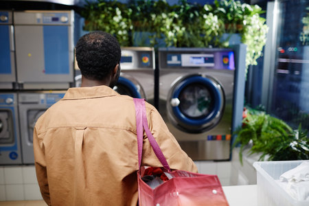 Rear view of young African American man in beige shirt carrying big bag with clothes on his shoulder...