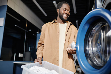 Happy young man in beige shirt opening door of washing machine before putting there clothes from basket while standing in front of cameraの写真素材