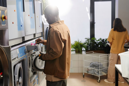 Young African American man putting washing powder into special small compartment in automatic machine in laundry cafeの写真素材