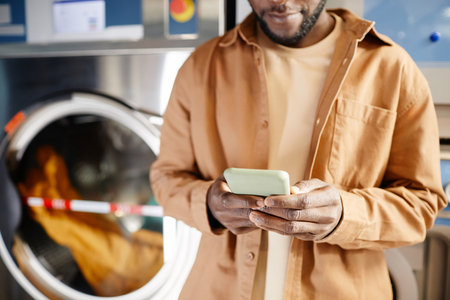 Hands of African American guy texting or watching online video in mobile phone while standing against automatic washing machineの写真素材