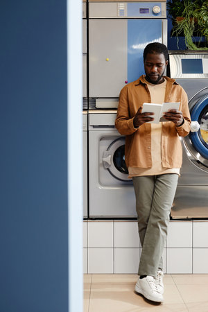 Young African American man in casualwear standing by washing machine and reading book in white cover in modern laundry cafeの写真素材