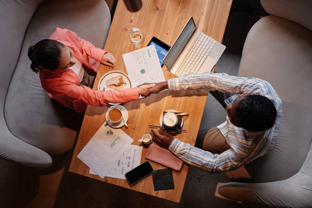 Above view of two successful white collar workers handshaking over table with mobile gadgets, financial documents, coffee and snackの写真素材