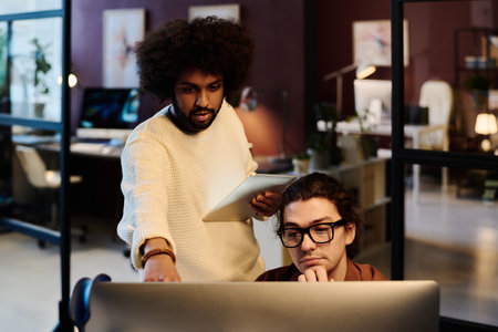 Young confident businessman with tablet explaining his colleague data on computer screen during discussion of project points at meetingの写真素材