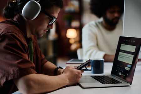 Serious young man with mobile phone texting in front of laptop with AI design tasks on screen while sitting by workplace against colleagueの写真素材